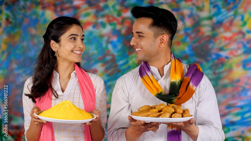 Indian youngsters with a plate of yellow Gulal and traditional Gujia on Holi - an auspicious occasion  Indian festival of colors  Indian siblings. A cheerful girl and a handsome guy wearing a white...
