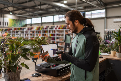 A man behind the cashier stand is scanning a price tag on a potted plant with a barcode reader.