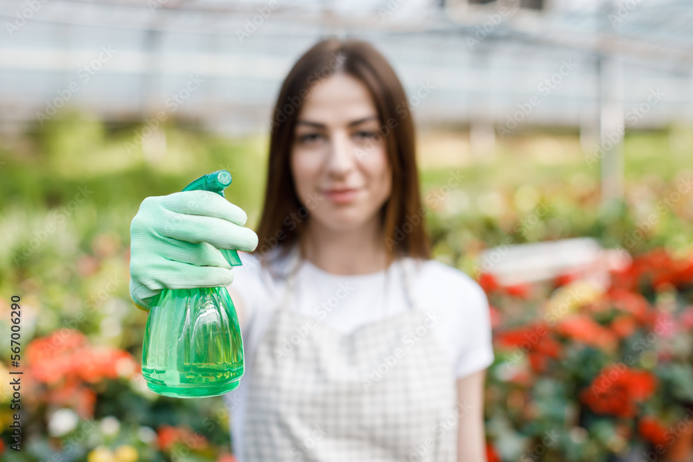 Gardening concept. Pretty smiling woman in apron holding sprayer in hand and looking to camera, standing in modern greenhouse on the background of plants and flowers