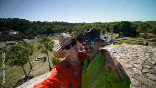 Mature woman and man couple wearing ethnic clothes, sunglasses, hats taking selfies at the top of Xcambo Mayan pyramid ruins in Mexico.