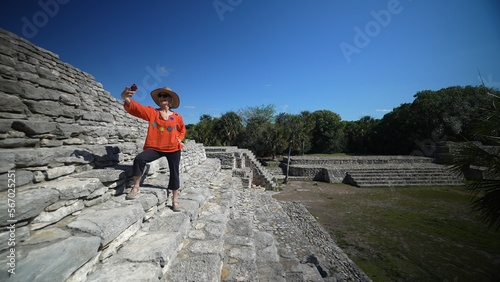 Mature woman wearing ethnic clothes, sunglasses, hat taking selfies with phone standing on stone steps of Xcambo Mayan pyramid in Yucatan Mexico. Concept of adventure is ageless.