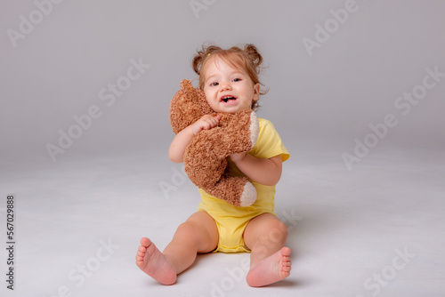 a baby girl in a yellow bodysuit sits and plays with a teddy bear on a white background