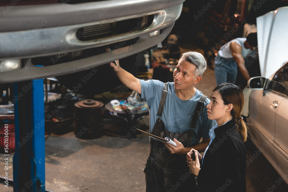 Senior male mechanic in uniform checking and diagnostic car while talking to young woman in business clothing with folding arms and informing about vehicle issue in garage while working Senior male mechanic in uniform checking and diagnostic car while talking to young woman in business clothing with folding arms and informing about vehicle issue in garage while working