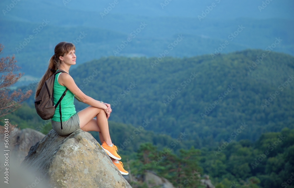 Naklejka premium Sportive female hiker sitting alone taking a break on hillside trail. Lonely woman enjoying view of evening nature from rocky cliff on wilderness path. Active lifestyle concept