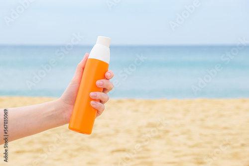 Womans hand holds a sunscreen spray in an orange bottle against the backdrop of a sandy beach and sea. The concept of applying sunscreen during a beach holiday.