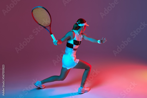 Back view of professional tennis player playing tennis over pink-purple background in neon light. Concept of sport, health, strength, action, motion, lifestyle.