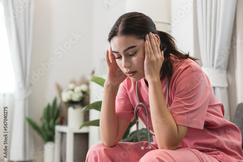 Upset female nurse sitting on a couch at home. Healthcare worker having headache. Doctors face heavy levels of stress. Young Caucasian caregiver in pink scrubs sitting hand touching temple tired face.