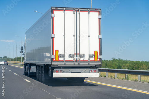 Rear view of white van semi truck drive on suburban asphalted highway road at summer day against blue sky, cargo transportation concept.