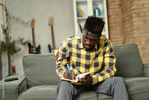 Young African man writing down notes while sitting on sofa at home..