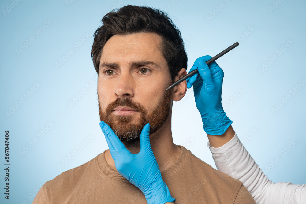 Aesthetic cosmetology concept. Man getting pencil marks on face skin for cosmetic surgery, blue background Aesthetic cosmetology concept. Man getting pencil marks on face skin for cosmetic surgery, blue background