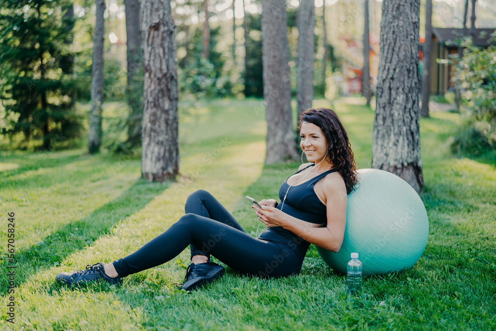 Relaxed sporty young female model takes break after aerobics exercises with fitness ball, sits on green grass, uses mobile phone, chats online, listens music in earphones, has athletic body. Relaxed sporty young female model takes break after aerobics exercises with fitness ball, sits on green grass, uses mobile phone, chats online, listens music in earphones, has athletic body.