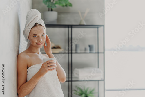 Pleased attractive woman has natural beauty  glowing smooth healthy skin  touches face  enjoys spa procedures  wears white bath towel on head and around body  drinks tea  poses in cozy bathroom