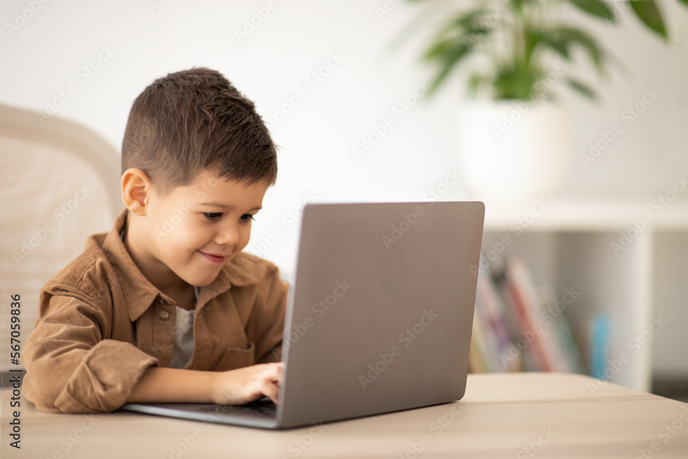 Cheerful small european boy sitting at table, typing on computer, play game, watch video in room interior Cheerful small european boy sitting at table, typing on computer, play game, watch video in room interior