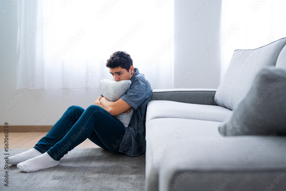 Pensive sad hispanic teenager boy sitting on floor and hugging pillow. Depression, anxiety and mental health concept Pensive sad hispanic teenager boy sitting on floor and hugging pillow. Depression, anxiety and mental health concept