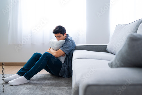 Pensive sad hispanic teenager boy sitting on floor and hugging pillow. Depression, anxiety and mental health concept © Egoitz
