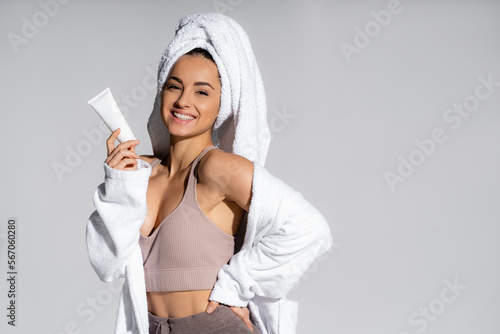 Cheerful young woman in towel and bathrobe holding cosmetic lotion isolated on grey. photo
