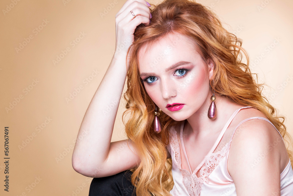 Beautiful red-haired girl with wavy hair, make-up, in a silk top and long earrings posing on a beige background in the studio. woman looking at camera Beautiful red-haired girl with wavy hair, make-up, in a silk top and long earrings posing on a beige background in the studio. woman looking at camera
