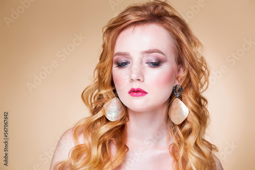 Beautiful red-haired girl with wavy hair and makeup posing on a beige background in the studio in large gold earrings