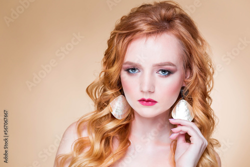 A beautiful red-haired girl with wavy hair and makeup poses on a beige background in the studio in round gold earrings. Woman touching earring and looking at camera