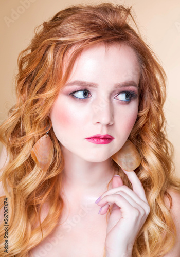 A beautiful red-haired girl with wavy hair and makeup poses on a beige background in the studio in round gold earrings. Woman touching an earring and looking away