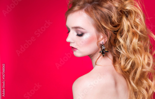 Beautiful red-haired girl with wavy hair and makeup posing on a red background in the studio in handmade earrings.