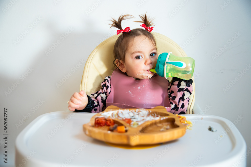 Portrait of cute toddler caucasian brunette girl with two tails eating by herself from modern wooden dish and drinking from green baby learning cup; self feeding concept, blw Portrait of cute toddler caucasian brunette girl with two tails eating by herself from modern wooden dish and drinking from green baby learning cup; self feeding concept, blw