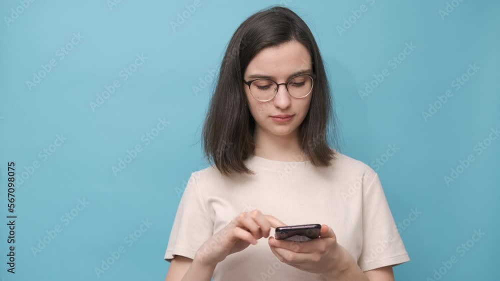 Portrait of a student girl with glasses and a beige blouse, holding a smartphone, looking for something on the phone, chatting with friends on a blue background. Online shopping, online training. Portrait of a student girl with glasses and a beige blouse, holding a smartphone, looking for something on the phone, chatting with friends on a blue background. Online shopping, online training.