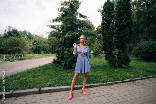 A beautiful girl with blond hair and long legs, in a short sexy summer purple skirt and shirt, with chic makeup and a stylish hairstyle, smokes on a path in the park.