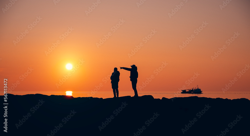 Silhouettes of two male friends looking together at a romantic sunset by the sea.