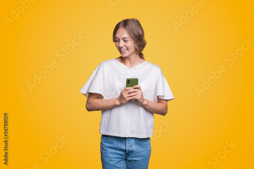 Laughing attractive positive young woman wearing casual stylish outfit sending text messages via mobile phone isolated over yellow background.