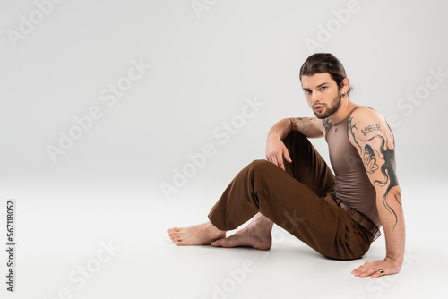 Full length of tattooed man looking at camera while sitting on grey background.