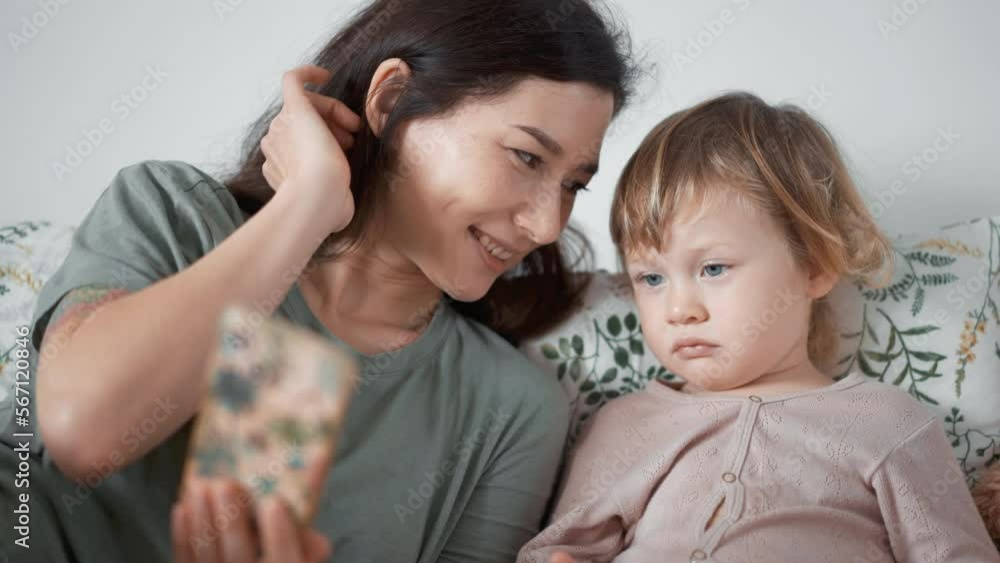 Asian young mother and curly boy daughter toddler kid look at phone screen video chat online. Woman and baby taking selfie or watch cartoons. Spend time together. Parenthood concept. bedroom morning Asian young mother and curly boy daughter toddler kid look at phone screen video chat online. Woman and baby taking selfie or watch cartoons. Spend time together. Parenthood concept. bedroom morning