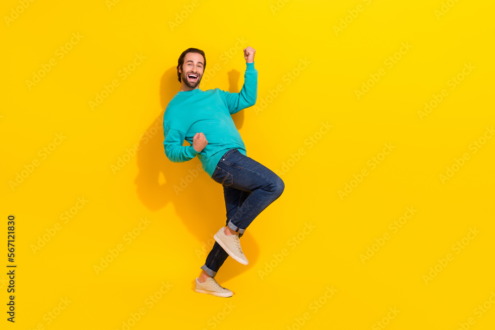 Full length photo of excited lucky man wear turquoise clothes rising fists empty space isolated yellow color background Full length photo of excited lucky man wear turquoise clothes rising fists empty space isolated yellow color background