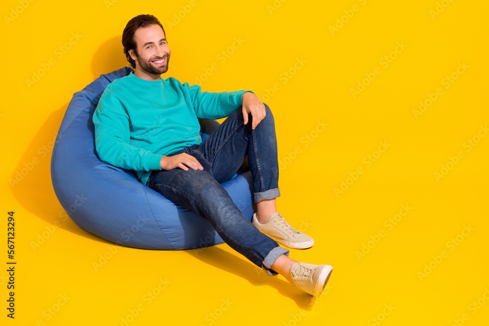 Full length photo of cheerful sweet man wear turquoise clothes sitting beanbag empty space isolated yellow color background Full length photo of cheerful sweet man wear turquoise clothes sitting beanbag empty space isolated yellow color background