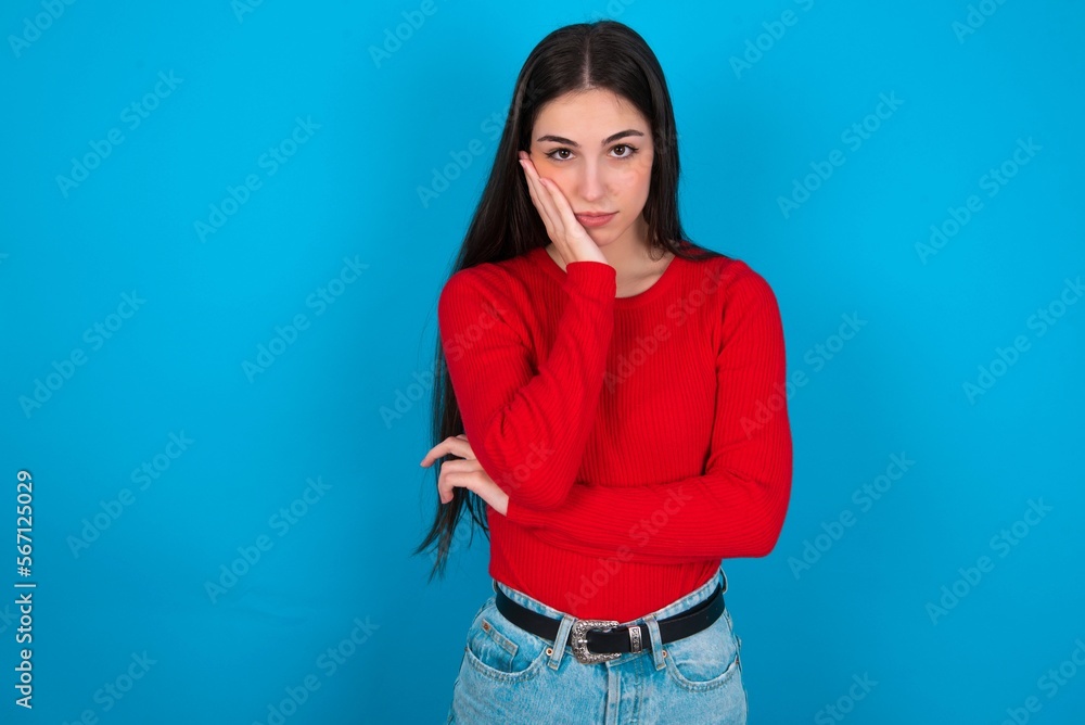 Very bored young brunette girl wearing red T-shirt against blue wall holding hand on cheek while support it with another crossed hand, looking tired and sick, Very bored young brunette girl wearing red T-shirt against blue wall holding hand on cheek while support it with another crossed hand, looking tired and sick,