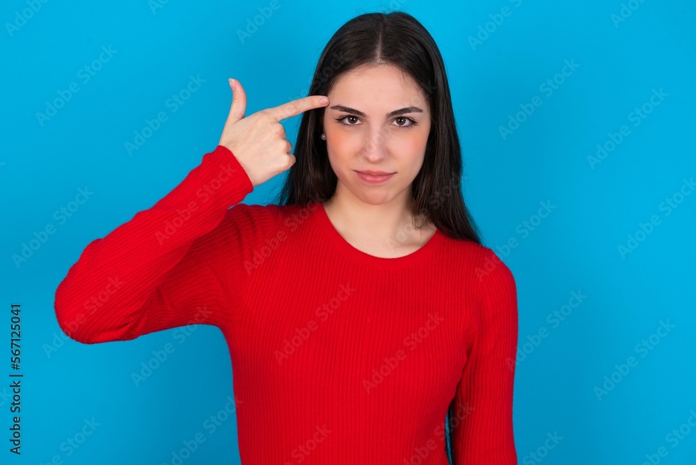 Unhappy young brunette girl wearing red T-shirt against blue wall makes suicide gesture and imitates gun with hand, curves lips keeps two fingers on temple, shoots, being tired of everything, Unhappy young brunette girl wearing red T-shirt against blue wall makes suicide gesture and imitates gun with hand, curves lips keeps two fingers on temple, shoots, being tired of everything,