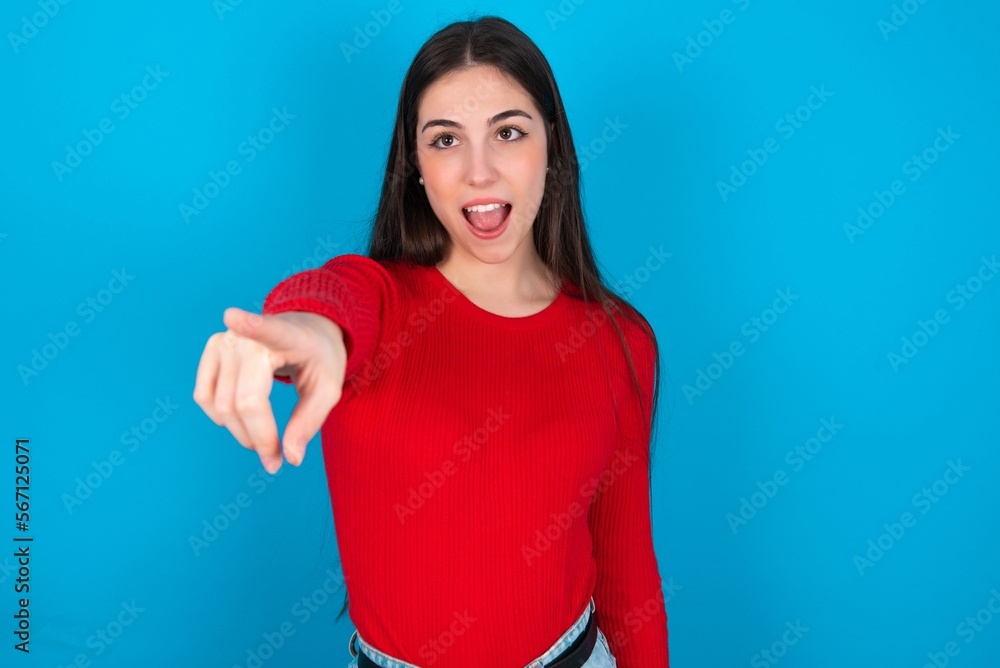 young brunette girl wearing red T-shirt against blue wall Pointing with finger surprised ahead, open mouth amazed expression, something on the front. young brunette girl wearing red T-shirt against blue wall Pointing with finger surprised ahead, open mouth amazed expression, something on the front.