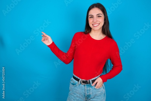 young brunette girl wearing red T-shirt against blue wall looking at camera indicating finger empty space sales