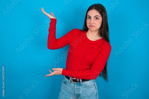 young brunette girl wearing red T-shirt against blue wall pointing aside with both hands showing something strange and saying: I don't know what is this. Advertisement concept.