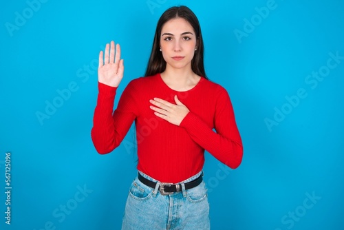 young brunette girl wearing red T-shirt against blue wall Swearing with hand on chest and open palm, making a loyalty promise oath