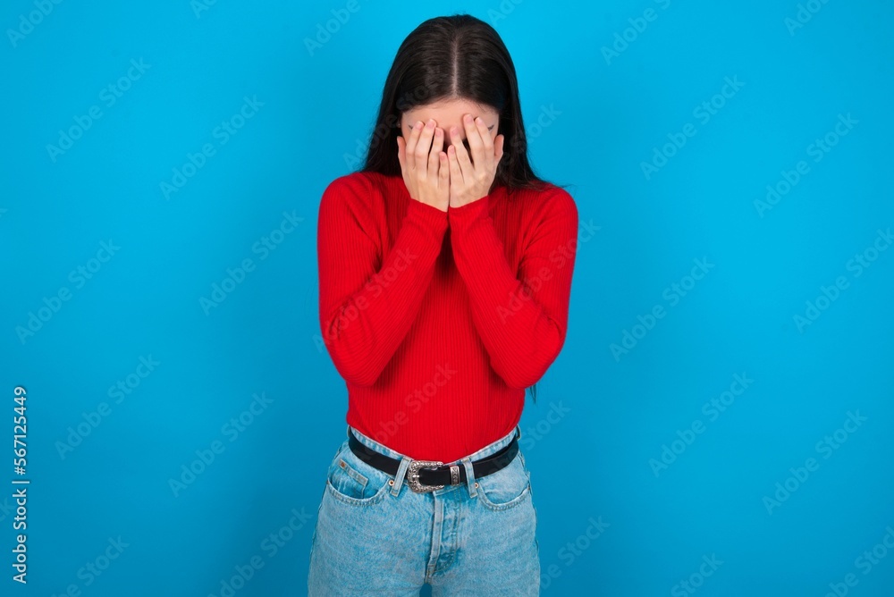 young brunette girl wearing red T-shirt against blue wall covering her face with her hands, being devastated and crying. Sad concept young brunette girl wearing red T-shirt against blue wall covering her face with her hands, being devastated and crying. Sad concept
