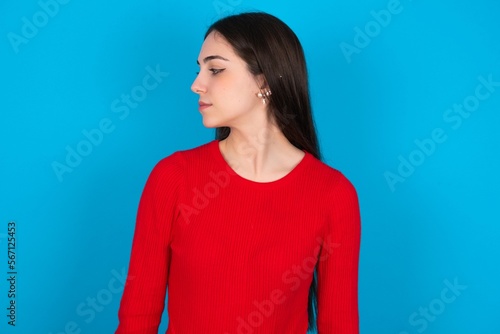 Close up side profile photo young brunette girl wearing red T-shirt against blue wall not smiling attentive listen concentrated
