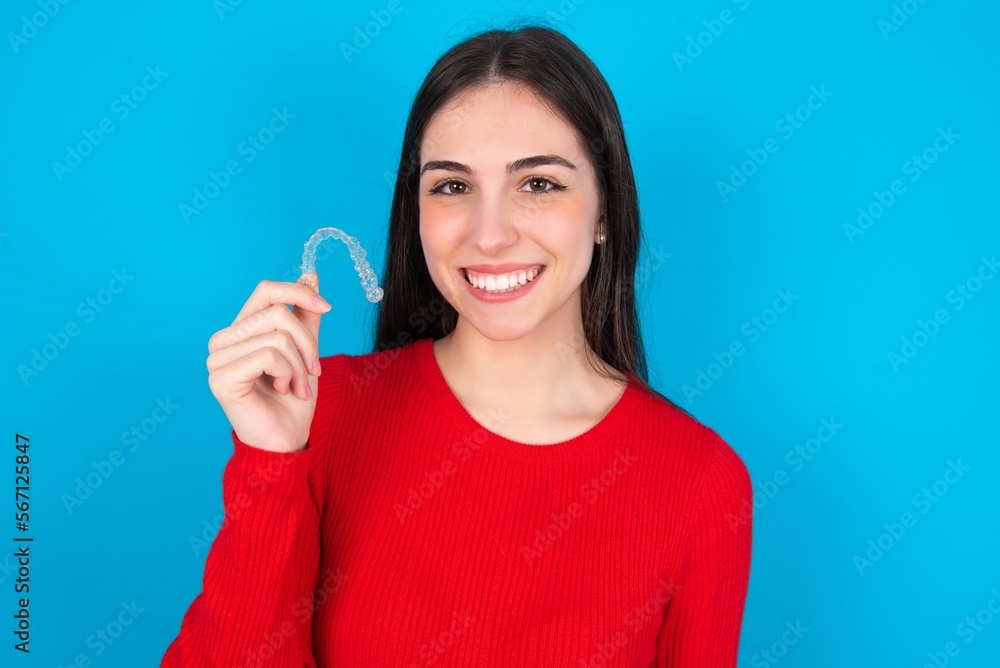 young brunette girl wearing red T-shirt against blue wall holding an invisible braces aligner, recommending this new treatment. Dental healthcare concept. young brunette girl wearing red T-shirt against blue wall holding an invisible braces aligner, recommending this new treatment. Dental healthcare concept.