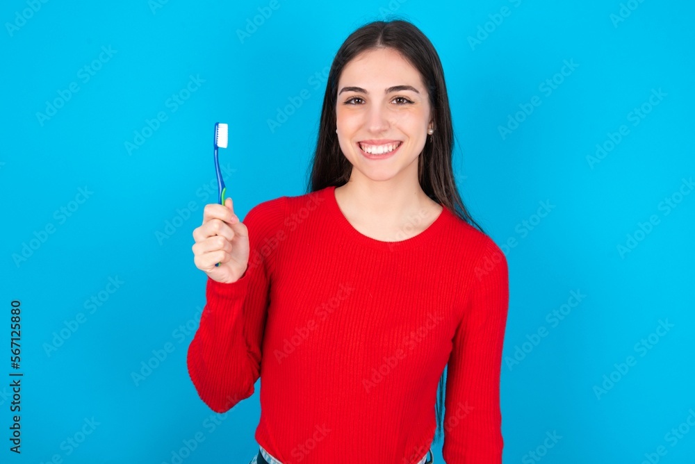 young brunette girl wearing red T-shirt against blue wall holding a toothbrush and smiling. Dental healthcare concept. young brunette girl wearing red T-shirt against blue wall holding a toothbrush and smiling. Dental healthcare concept.