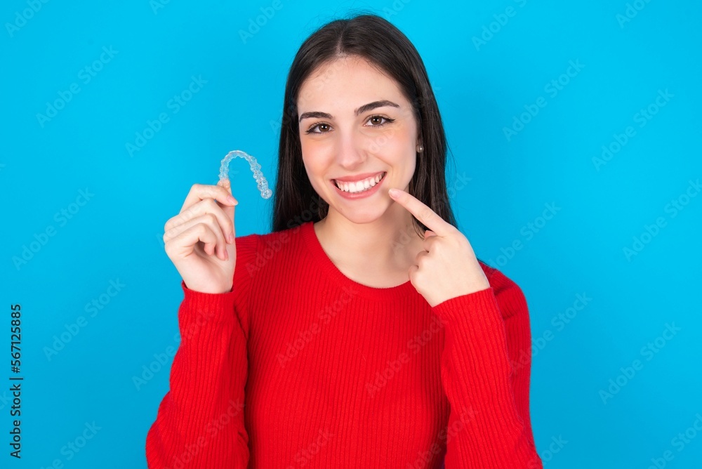 young brunette girl wearing red T-shirt against blue wall holding an invisible aligner and pointing perfect straight teeth. Dental healthcare and confidence concept. young brunette girl wearing red T-shirt against blue wall holding an invisible aligner and pointing perfect straight teeth. Dental healthcare and confidence concept.