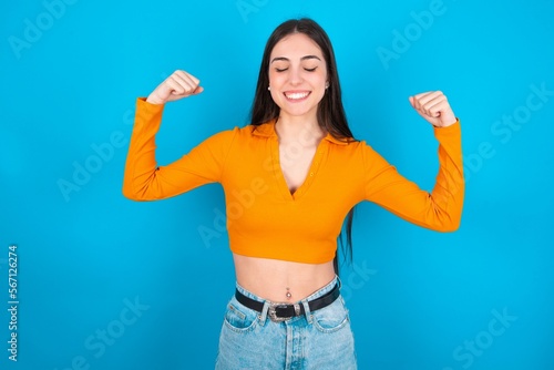 Strong powerful young caucasian brunette girl wearing orange crop top against blue wall toothy smile, raises arms and shows biceps. Look at my muscles!