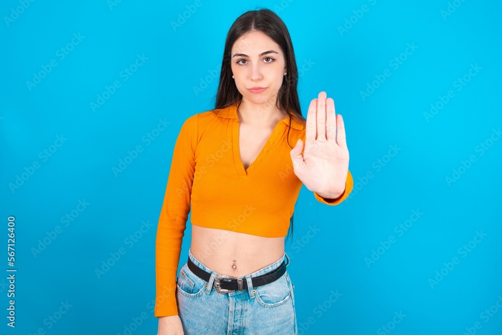 young caucasian brunette girl wearing orange crop top against blue wall shows stop sign prohibition symbol keeps palm forward to camera with strict expression