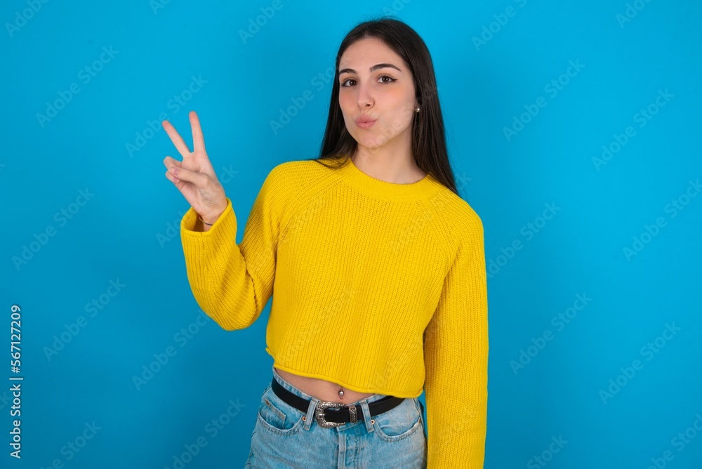 young brunette girl wearing yellow knitted sweater against blue wall makes peace gesture keeps lips folded shows v sign. Body language concept young brunette girl wearing yellow knitted sweater against blue wall makes peace gesture keeps lips folded shows v sign. Body language concept