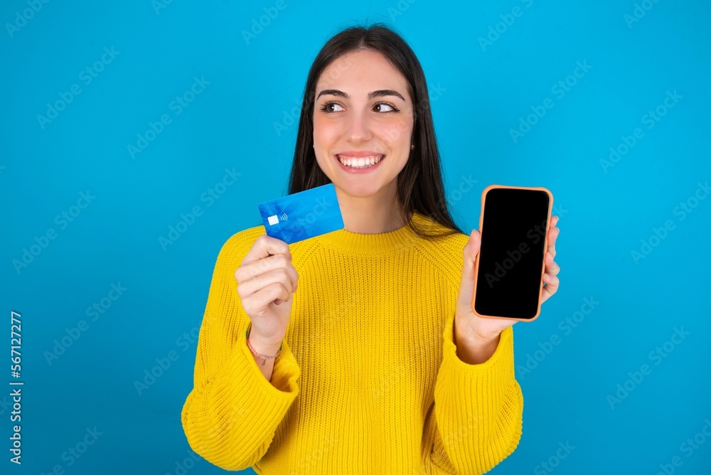 young brunette girl wearing yellow knitted sweater against blue wall holding bank card modern device looking empty space young brunette girl wearing yellow knitted sweater against blue wall holding bank card modern device looking empty space