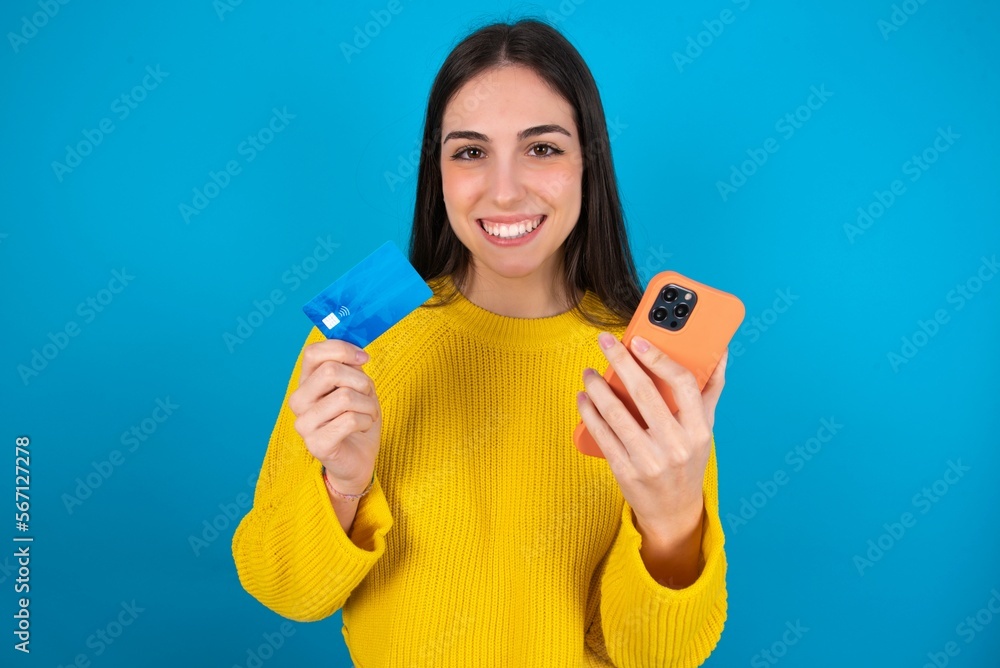 young brunette girl wearing yellow knitted sweater against blue wall wears stereo headphones on ears holds modern mobile phone and credit card young brunette girl wearing yellow knitted sweater against blue wall wears stereo headphones on ears holds modern mobile phone and credit card